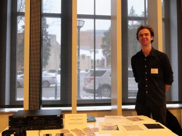 Latimer Rogland standing by their project, "Brokenness, Resolution, and Tactility Within Electronic Music and Physical Mediums," with a sound machine, headphones, and zines on a table.